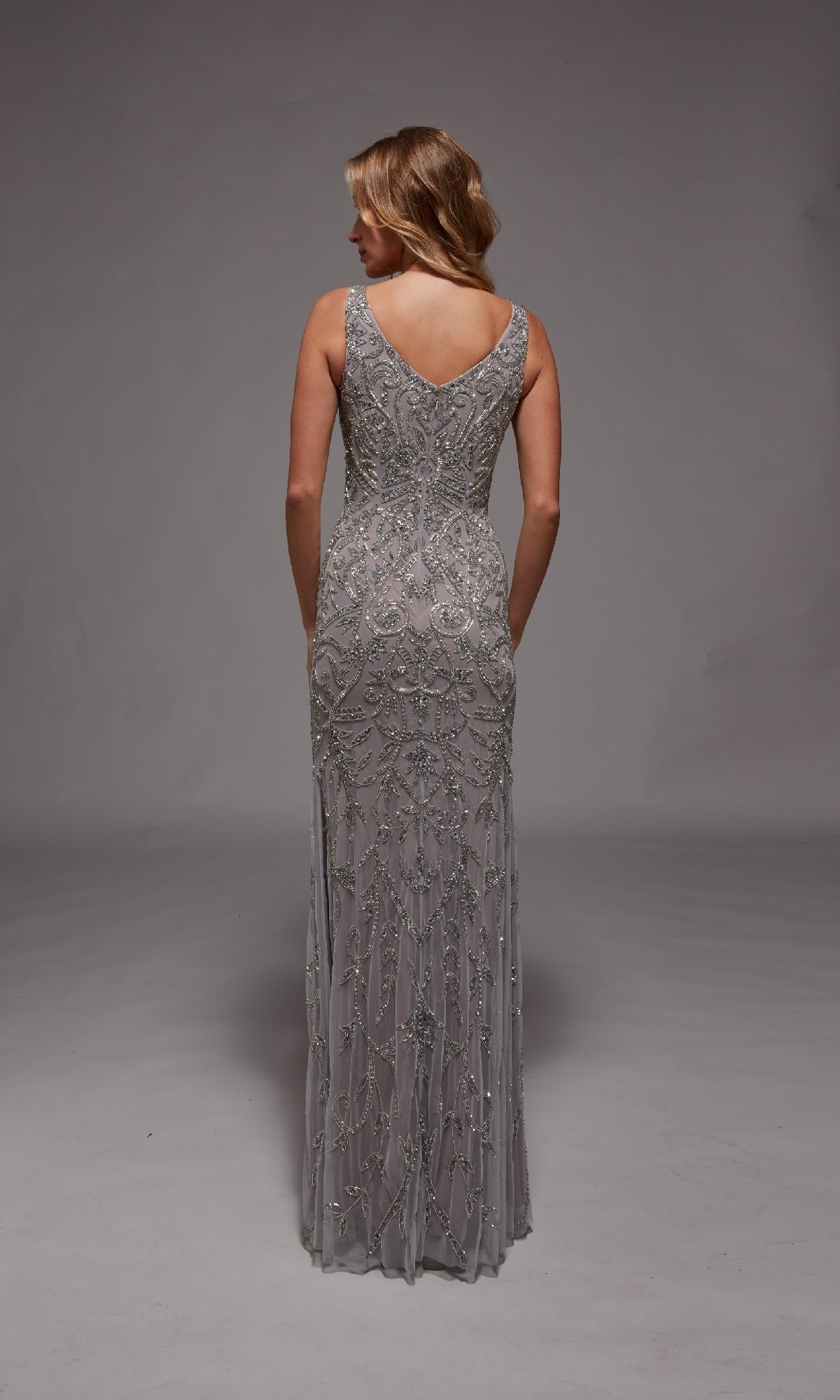 A woman stands facing away in the Alyce 27799, a sleeveless silver gown with intricate beadwork and a V-back—an elegant long formal dress ideal for any special occasion.